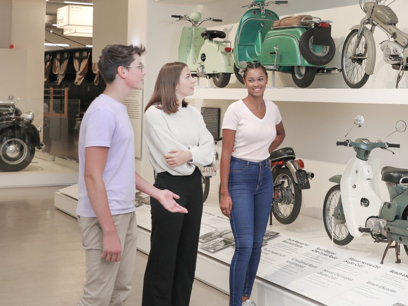 : A small group of teenagers look at various mopeds and motorcycles in the exhibition "Mobility".